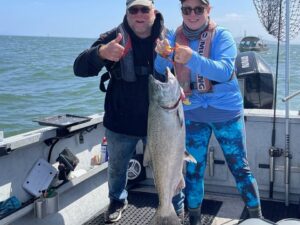 Smiling customer holds up a giant Chinook salmon caught at Buoy 10 while guide Marvin Henkel gives a thumbs-up sign.